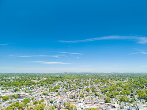 Aerial drone view over a sprawling residential suburban neighborhood on a clear sunny day, featuring numerous houses, green trees, and distant cityscape on the horizon