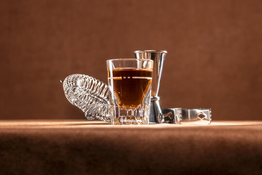 Shot glass of whiskey sitting in front of common stainless steel bartending tools like a strainer, a jigger, and ice tongs. 