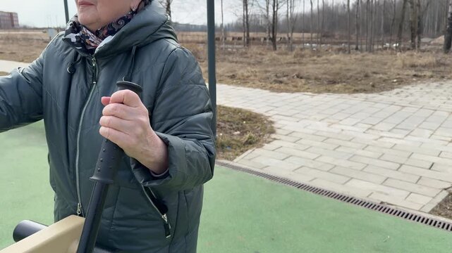 Senior woman exercising on elliptical machine in park playground on spring day, wearing jacket, hat and glasses, steady rhythm.