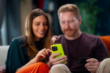 Fototapeta premium A man and a woman sit close on a couch. They look at a smartphone and share a credit card. They seem to talk and smile while shopping online in their living room.