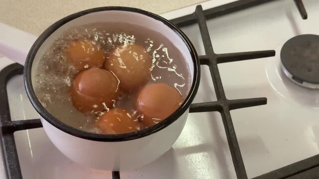 Close-up boiling brown eggs in ladle on gas stove, white enamel pot filled with bubbling water, gentle steam rising, homely morning.