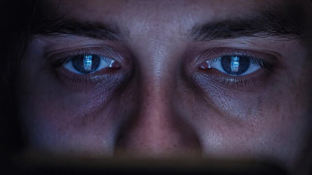 Extreme close up of human eyes reflecting social media feed continuously scrolling on smartphone screen in dark room at night.