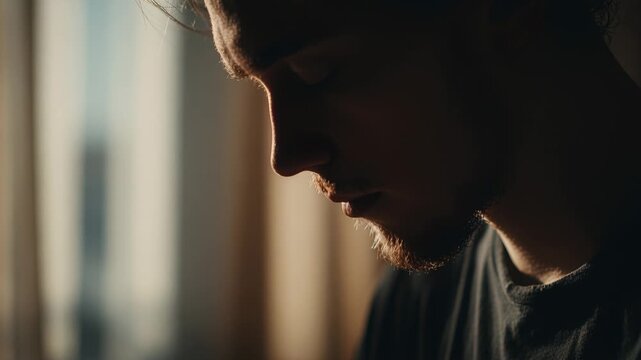 Young bearded man bows head silhouetted against warm window backlight