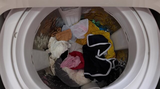 A top-loading washing machine with the lid open, showing a mixed load of laundry inside the drum. The machine appears moderately filled and ready for a wash 