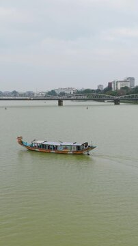 Vertical screen Bright Wooden Craft Navigating River. Vibrant Wooden Boat Captured Near Riverside Bridge And Quay. Brightly Painted Wooden Boat With Canopy Maneuvering Close To Riverside