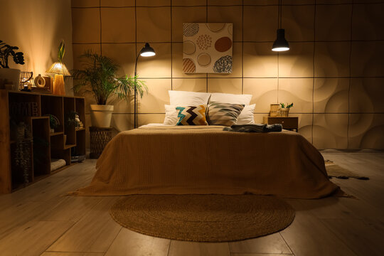 Interior of cozy bedroom with blankets on bed, shelf unit and glowing lamps at night