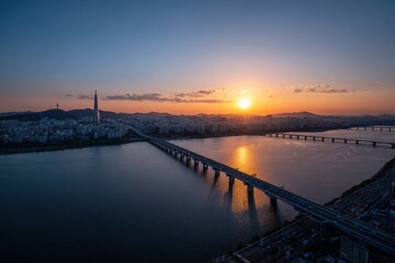 Fototapeta premium Golden Hour Sunset Over a Bustling Cityscape with a Long Bridge Spanning a Wide River Reflecting Warm Light and Silhouetted Buildings in the Distance