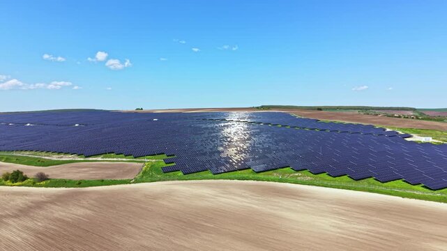 A wide view shows many solar panels arranged in rows across a large field. The sun shines bright, and the land is mainly brown with some green patches.
