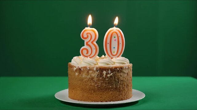 A small birthday cake with brightly lit number 30 candles stands prominently on a white plate against a vibrant green background, celebrating a significant milestone.