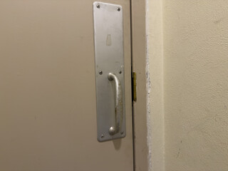 Fototapeta premium Close-up of a textured beige utility door with a worn silver metal pull handle, showcasing industrial hardware, visible screws, peeling paint, a distressed door frame, and a rough plastered wall.