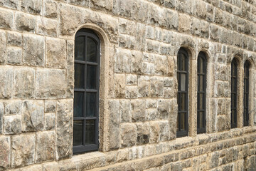 Fototapeta premium black arched old windows on long grey stone wall, building facade with uneven chiseled blocks, side angle view, ancient architecture concept