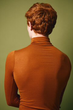 Redheaded man with short curly hair wearing tight orange turtleneck sweater standing with back to camera against green background indoors.