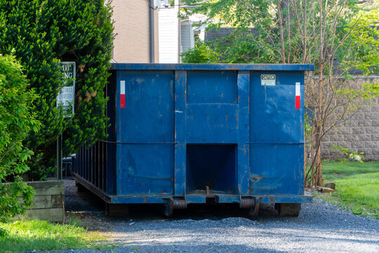 Large weathered blue roll-off dumpster on a gravel driveway, surrounded by vibrant green foliage and residential buildings, signifying construction, renovation, or waste removal in a suburban setting.