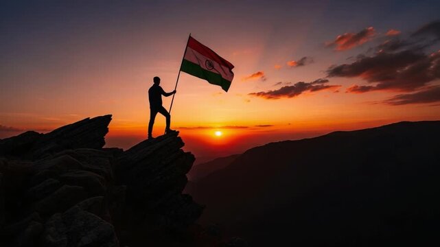 Silhouette of a young man standing on a mountain peak holding a flag at sunset representing victory and achievement