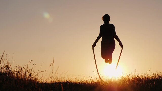 Silhouette of a woman jumping rope at sunset in a field full of grass, fitness and healthy lifestyle motion.