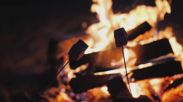 Two marshmallows on sticks gently rotate over a brightly burning campfire at night, creating a cozy and warm outdoor scene.