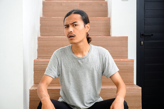Young Indonesian man with long hair sitting on wooden stairs with a confused or questioning expression on his face portraying curiosity or disbelief