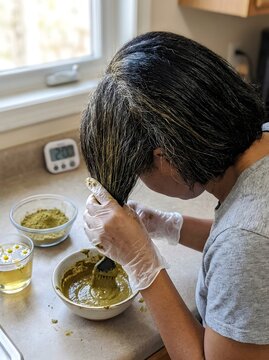 Woman with Gray Roots Applying Green Henna Paste on Hair with Brush and Gloves Next to Chamomile Tea Bowl and Kitchen Timer for Natural Hair Dye at Home