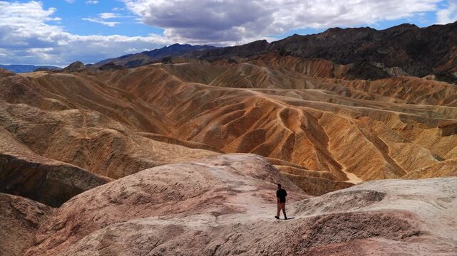 Lone hiker explores the vibrant eroded mudstone formations at Zabriskie Point in Death Valley National Park. Golden waves of sedimentary rock stretch to distant mountains under dramatic clouds.