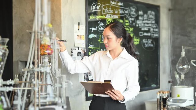 Young Asian Female Scientist Taking Notes on Clipboard in Chemistry Laboratory with Glassware and Chalkboard