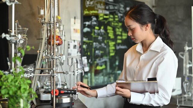 Young Asian Female Scientist Taking Notes on Clipboard in Chemistry Laboratory with Glassware and Chalkboard