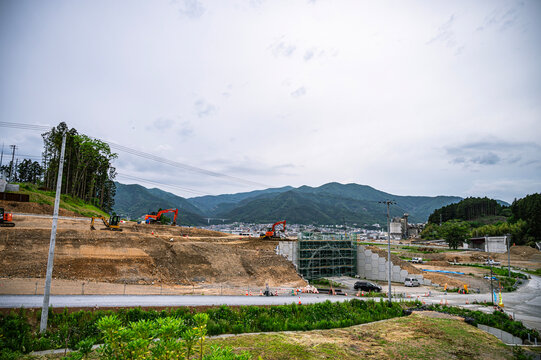 山を背景に複数の重機が稼働する、大規模な道路・トンネル建設工事現場の全景風景。 / Panoramic view of a large-scale road and tunnel construction site with heavy machinery and mountains.