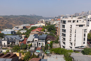 Obraz premium Sunlit urban residential neighborhood with mixed traditional houses and a modern apartment building on a hillside, showing rooftops, narrow streets, greenery and a city skyline of Yerevan, Armenia