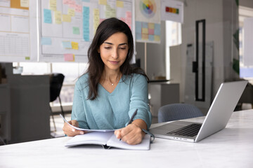 Young professional woman working in modern office, writes in notebook while holds documents,...