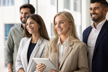 Obraz premium Group of four diverse business professionals, male and female colleagues dressed in elegant business suits standing together in modern office, smile, looking aside. Teamwork, leadership, joint success