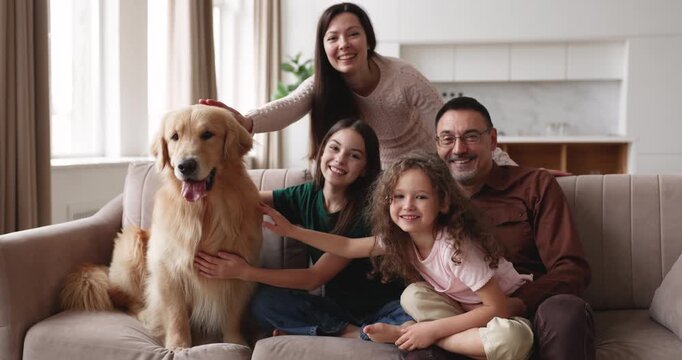 Smiling Caucasian couple with two daughters and golden retriever dog posing for portrait sitting on couch in living room, showing family love, feeling bonding, enjoying leisure time together at home
