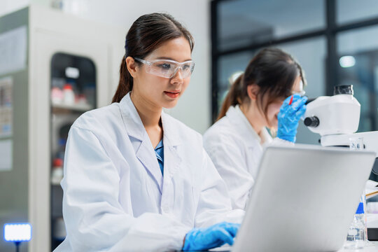 Asian female scientists team using laptop and microscope in modern laboratory, clinical research specialists working on scientific experiment, medical biotechnology analysis, pharmaceutical.
