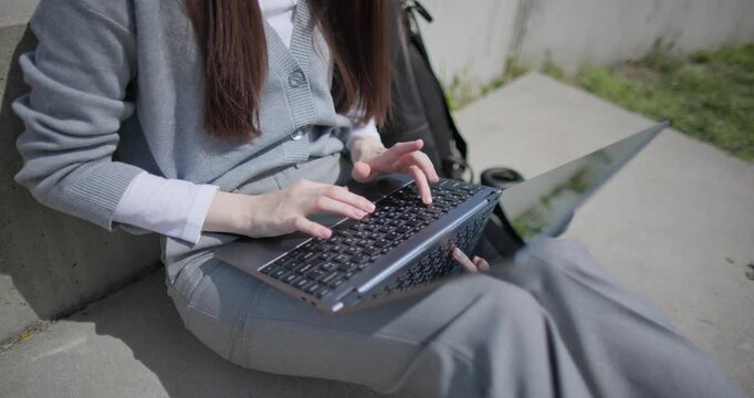 A hand-held shot of a young female student in casual clothes and glasses working and typing on a laptop while sitting in an urban park - close-up, moving the camera from her hands to her face