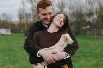 Fototapeta premium A joyful couple outdoors, embracing a small goat kid in a grassy field, wearing cozy sweaters. A candid lifestyle moment highlighting affection, companionship, and rural life.