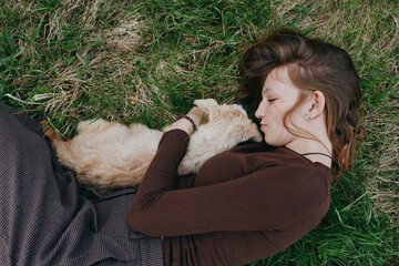 Fototapeta premium Woman lies on grass cuddling a dog, cozy outdoor moment in a brown sweater, natural light, relaxed lifestyle scene, affectionate bond, peaceful mood and quiet countryside feeling.