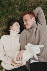 Fototapeta premium Smiling couple lying on grass in cozy turtlenecks, holding a fluffy goat kid between them. Warm natural light conveys relaxed lifestyle, gentle affection, and countryside charm.