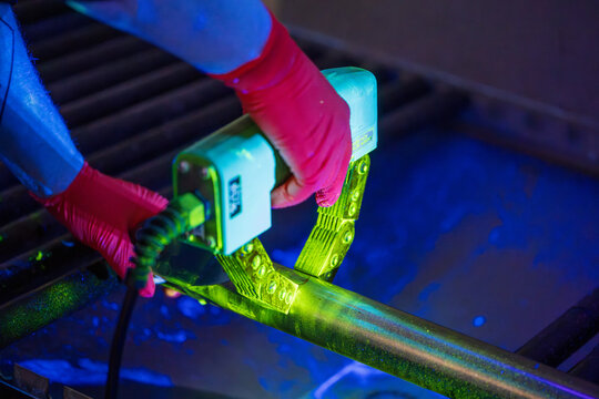 NDT Inspector in Red Gloves Using a Handheld AC Electromagnetic Yoke for Fluorescent Magnetic Particle Inspection MPI on a Metal Pipe. Professional Nondestructive Testing Under Vivid UV Light