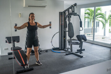 Fototapeta premium Young woman in black outfit performing dumbbell exercises with arms extended in a bright modern gym with large windows and equipment. Active fitness concept