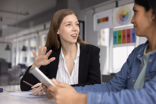 Young businesswoman in formal attire lead conversation with female colleague holding digital tablet at briefing in boardroom in modern office. Brainstorm, ideas exchange during creative team meeting