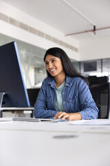 Plakat Smiling Indian female office employee working on computer seated at table in office, watching training video or online course on pc, reading documentation or work-related articles, using digital tools
