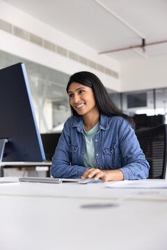 Smiling Indian female office employee working on computer seated at table in office, watching training video or online course on pc, reading documentation or work-related articles, using digital tools