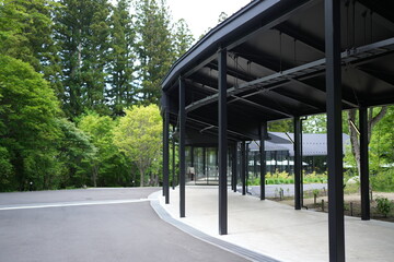 Fototapeta premium Modern pavilion stands beside green trees. Black steel columns support curved rooflines. Walking path curves gently beside the structure. Fresh leaves and tall pines surround the scene