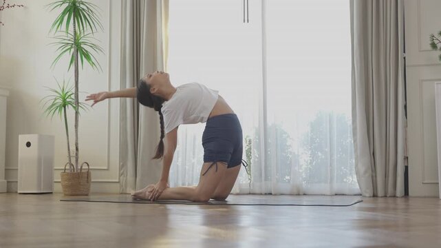 Peaceful woman stretching back doing yoga camel pose on floor mat inside bright home living room during morning exercise routine for healthy flexible active wellness fitness lifestyle