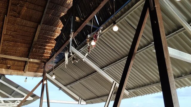 Low angle view of rustic open-air shelter ceiling featuring woven palm leaf thatch and corrugated metal roofing. Wooden support beams with hanging light bulbs in traditional tropical structure.