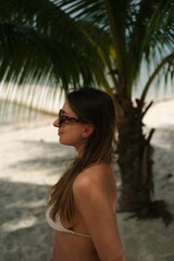 Obraz premium Woman in Bikini on Tropical Beach: A young woman with long brown hair and sunglasses, wearing a white bikini, stands on a sandy beach with a palm tree behind her.