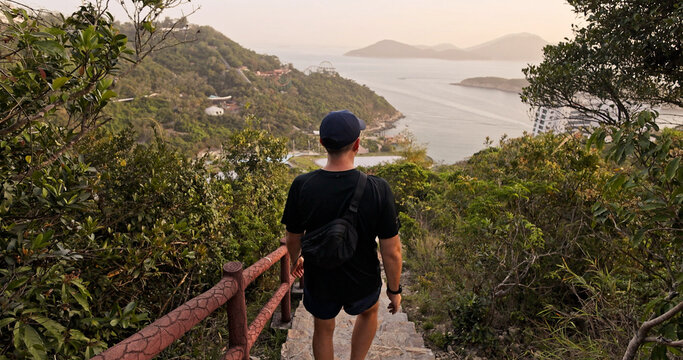 Back view of male hiker descending stone steps. Scenic Hong Kong island coastline and sea view. Lush green hills at sunset. Active lifestyle and travel concept.
