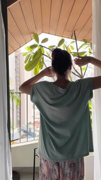 Cute woman in pajamas stands on a sunlit balcony among tropical plants, looking at the city and and stretching. Symbolizes morning routine and waking up. Ideal for lifestyle and wellness content