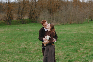Fototapeta premium Young couple embraces in a green field while holding a small baby goat as they share a tender kiss during a calm outdoor lifestyle moment.