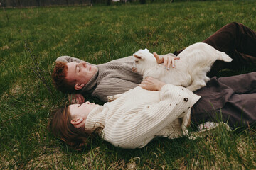 Fototapeta premium Couple and a playful goat kid lie together on a grassy field, sharing warm affection in soft daylight. A candid lifestyle moment showing relaxation, connection, and natural outdoor comfort.