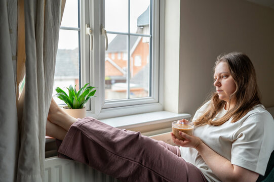 Exhausted woman sitting by window with coffee at home