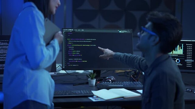 Male software engineer pointing at monitor while explaining code script to his female colleague in a modern office.